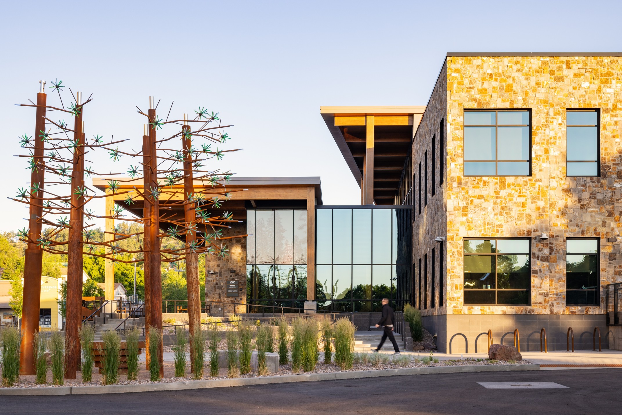Outside the Mountain Line Downtown Connection Center, the building has a glass entryway and a stone facade. There are short shrubs and tall spindly fake tress outside lining the street.