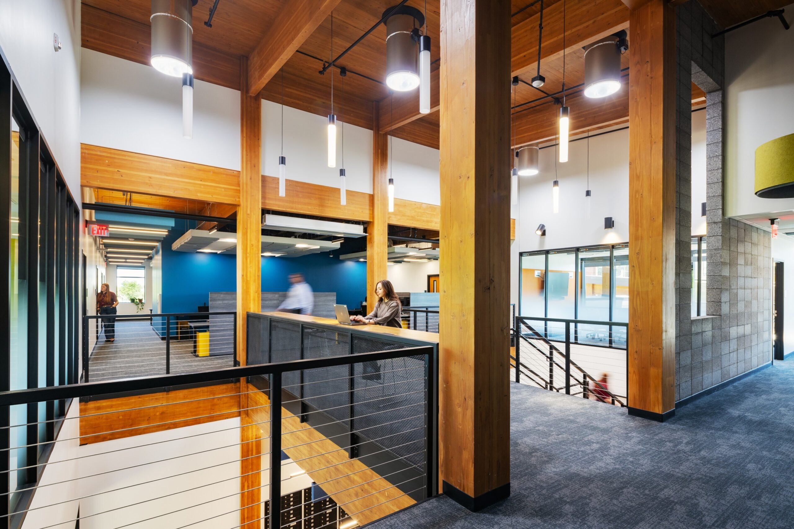 Inside the Mountain Line Downtown Connection Center, a tall-ceilingedecond floor of the building with light wells to the first floor and a staircase. One wall is bright blue and the others are white, tall wooden pillars support the wood beamed ceiling. Several people walk around.