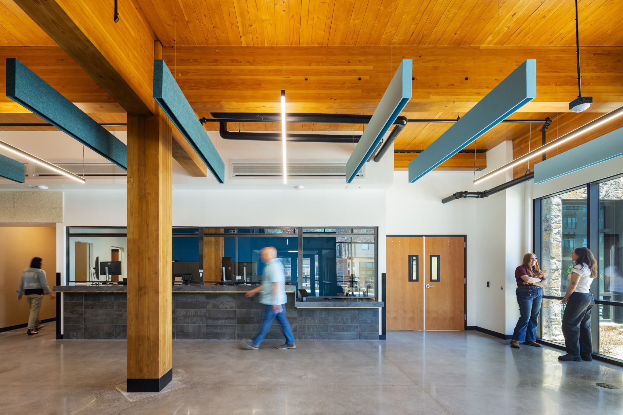 Inside the lobby of the Mountain Line Downtown Center - a light filled room with wooden ceilings, concrete floor, a check in desk, and several people walking through.