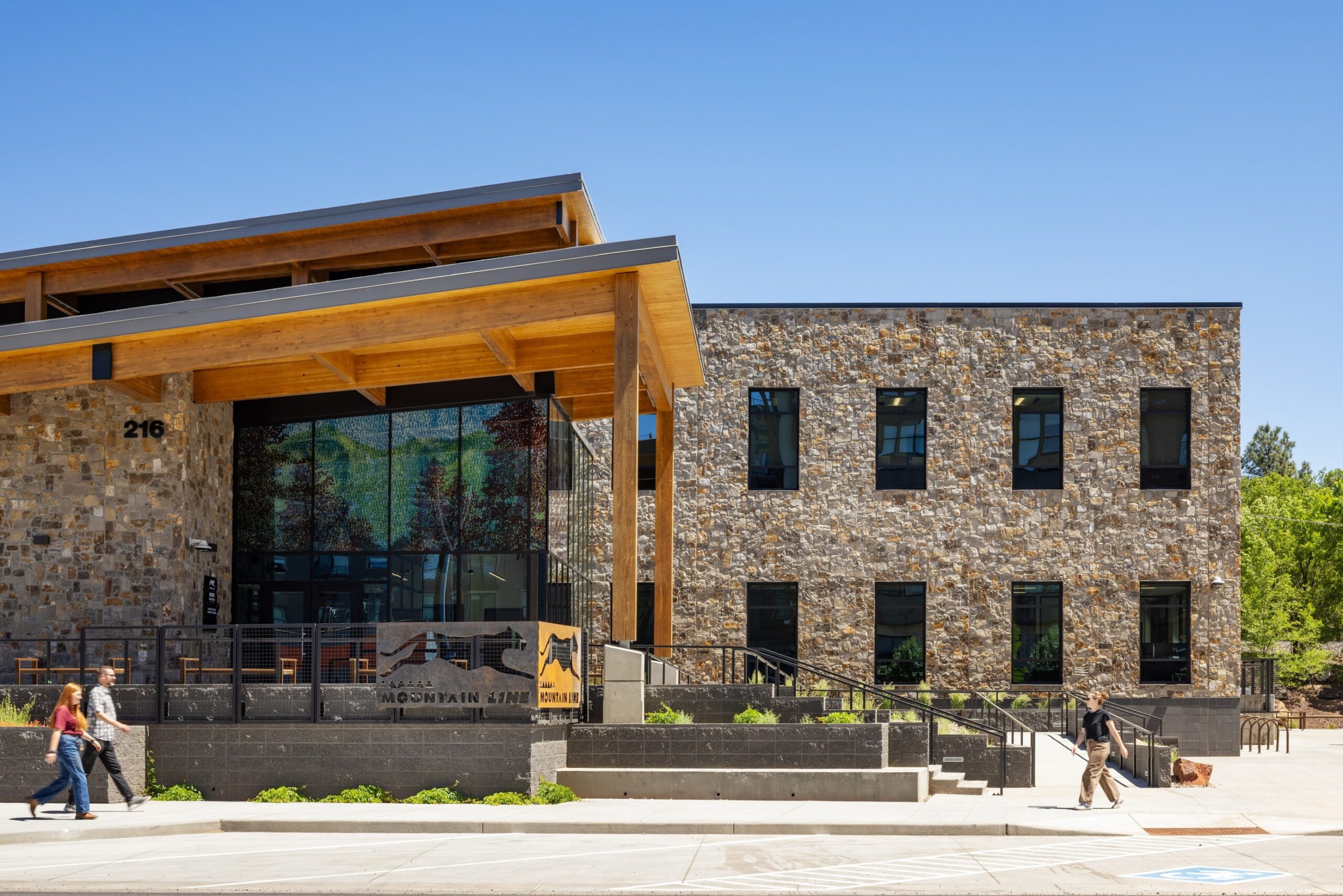 Outside of the Mountain Line Downtown Connection Center. A two story building with a glass entryway, slanted wooden roof, and stone facade. There are steps and plants leading up to the entrance, signage with a mountain lion, and blue skies behind the building.