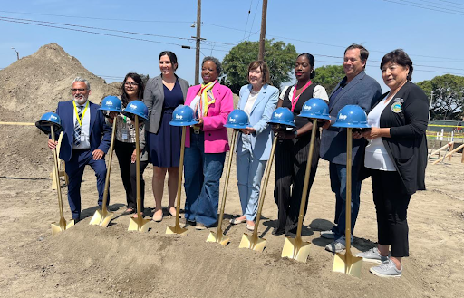 Eight people stand on a construction site holding golden shovels with blue helmets on top, posing for a groundbreaking ceremony celebrating a shovel ready brownfield project. Dirt piles and construction equipment are visible in the background.