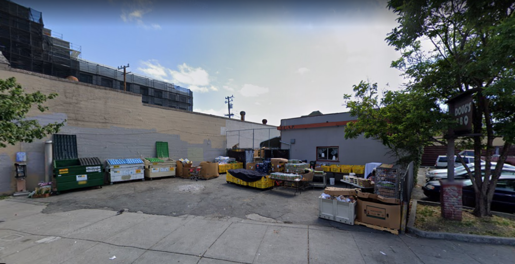 A small urban lot operating as a food bank in Emeryville, with dumpsters, stacked crates, and boxes next to a building. A tree and a parking sign are on the right, with cars partially visible. The sky is partly cloudy.