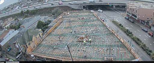 Wide view of a brownfield project construction site with workers on a large framework, next to busy streets and an elevated highway. Nearby are residential buildings, trees, and several cars driving on the roads.