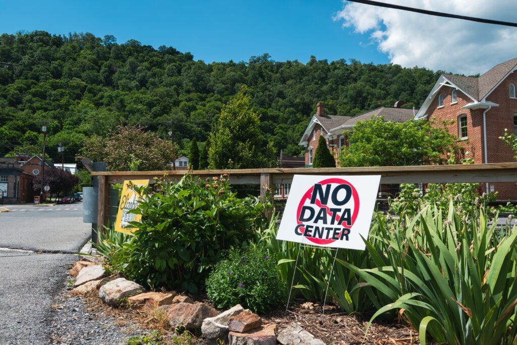A NO DATA CENTER yard sign is posted in a garden in a suburban neighborhood with green hills in the distance