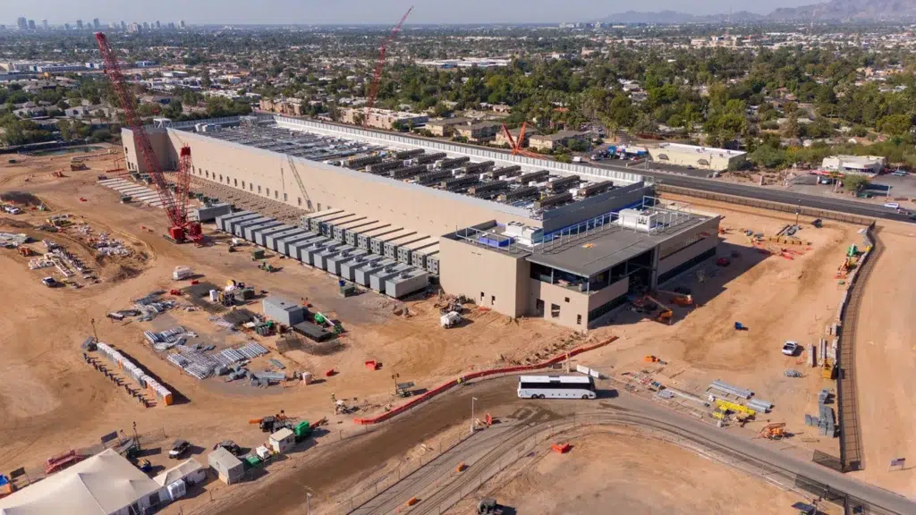Aerial view of a large construction site featuring a long, rectangular data center surrounded by equipment, vehicles, and dirt lots, with a homes and mountains in the distance. 